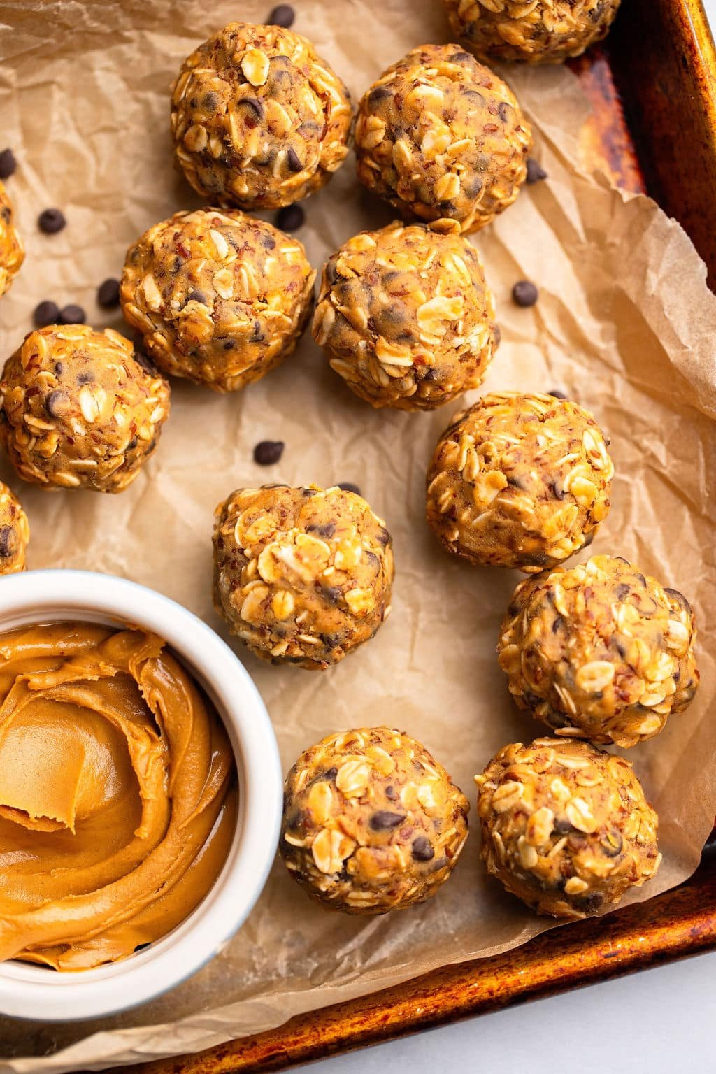 Near the peanut butter energy balls next to a small bowl of peanut butter in a baking sheet lined by parchment.