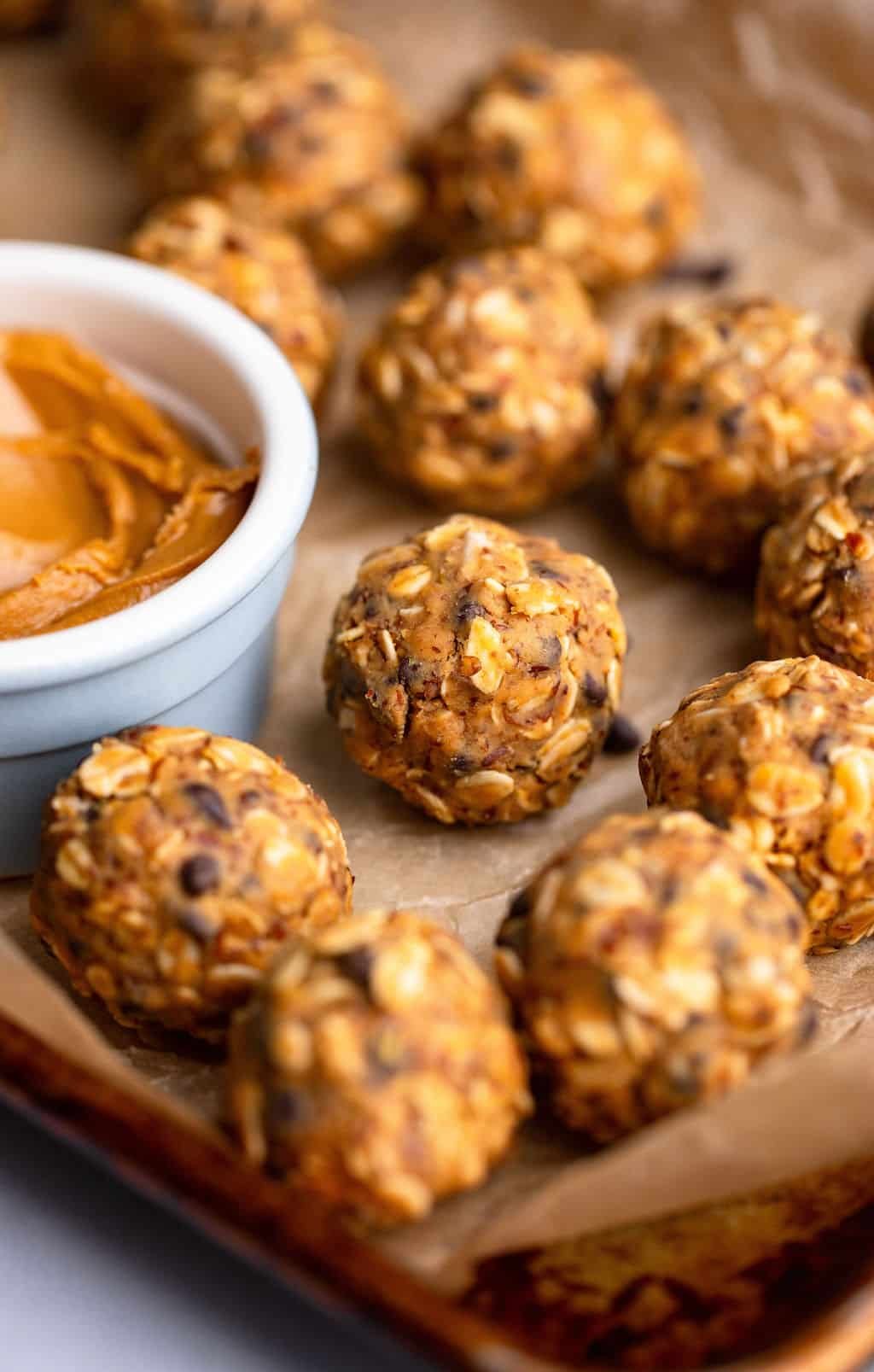 Near the peanut butter energy balls next to a small bowl of peanut butter in a baking sheet lined by parchment.